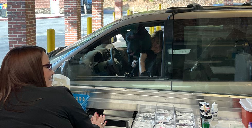 A person with a large black dog in a car at a pharmacy drive-thru window, interacting with a pharmacist.